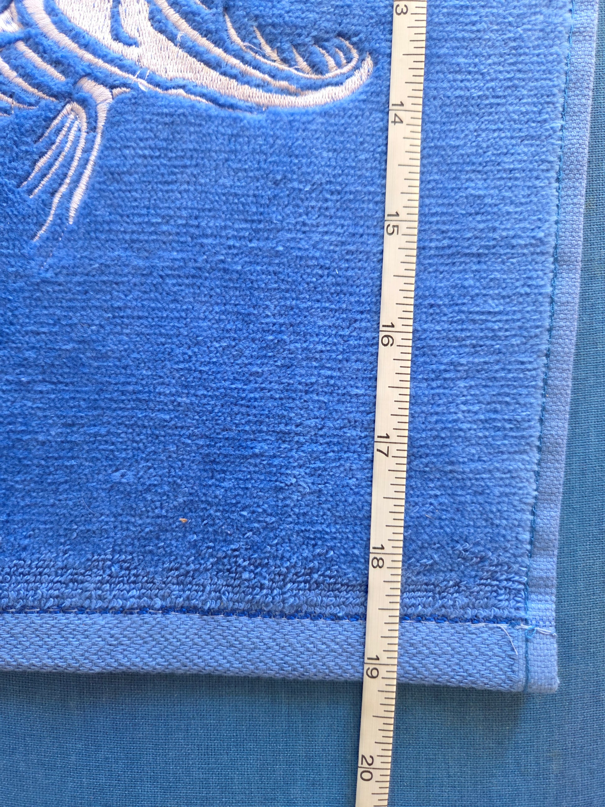 Blue towel with white embroidery on a blue background, measured using a ruler.
